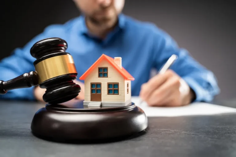 Miniature house model with a gavel in the foreground and a person signing documents in the background, representing property legal services.